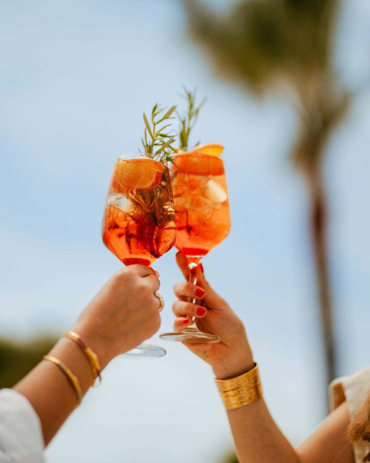 Two spritz glasses toasting against the blue sky and palm trees