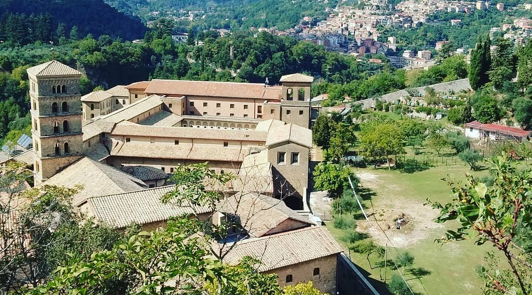 The Monastery of Santa Scolastica in Subiaco by day: the pale stone structure with its Romanesque mullioned bell tower, tiled roofs and cloister, overlooking the wooded valley