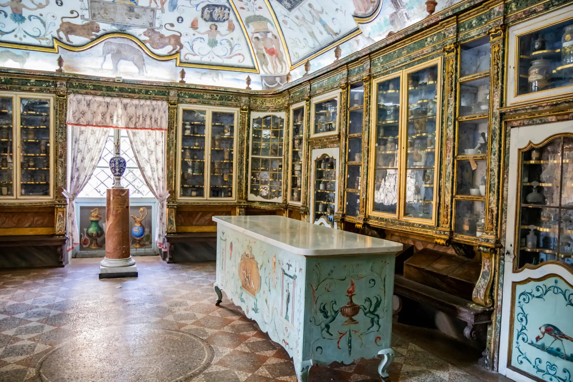 Interior of the ancient pharmacy of the Certosa di Trisulti with dark green and gilded furniture, cabinets of antique jars and stills, a marble counter and a trompe-l'œil frescoed ceiling