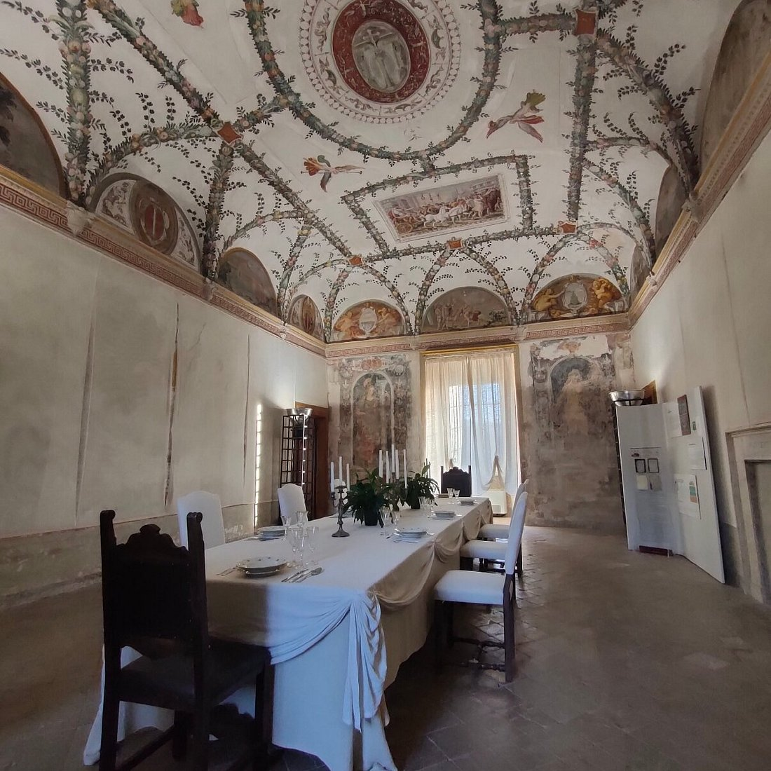 Interior hall of the Rocca Abbaziale in Subiaco with a frescoed cross-vaulted ceiling, a laid table and luminous curtains