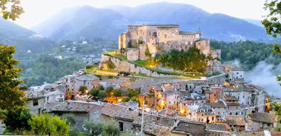 Panoramic view of the medieval village of Subiaco at dusk: the terraced town on the hill with the Rocca Abbaziale at the top, tiled roofs and mist in the valley