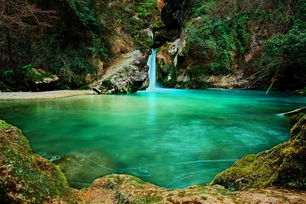 Emerald pool of the Lago di San Benedetto in Subiaco with a silky waterfall, moss-covered boulders and wild vegetation
